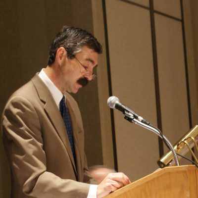 A man with a mustache standing at a podium.