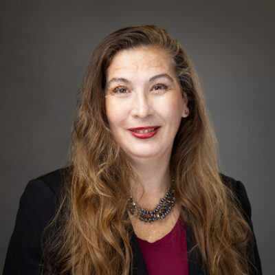 A smiling woman with long hair wearing a blazer and a necklace against a grey background.