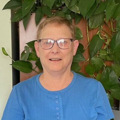 A woman with short hair and glasses is smiling and standing in front of a leafy green plant. She is wearing a blue shirt.