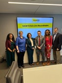 Six people standing in a conference room in front of a presentation screen displaying "Lived Crisis Line Broadcastthon.