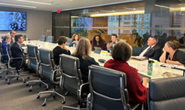 A group of people seated around a conference table in an office, having a meeting with a presentation screen in the background.