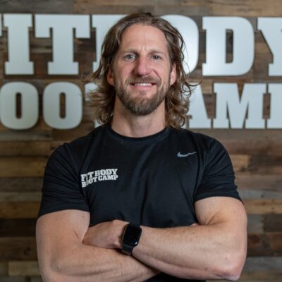 A person with long hair and a beard smiles while crossing their arms, wearing a "Fit Body Boot Camp" shirt. A wooden wall with a partial sign is in the background.