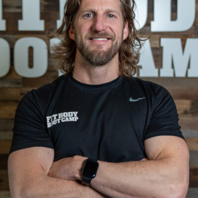 Man with long hair and beard wearing a black T-shirt and smartwatch, standing with arms crossed in front of a wooden wall with large white letters.