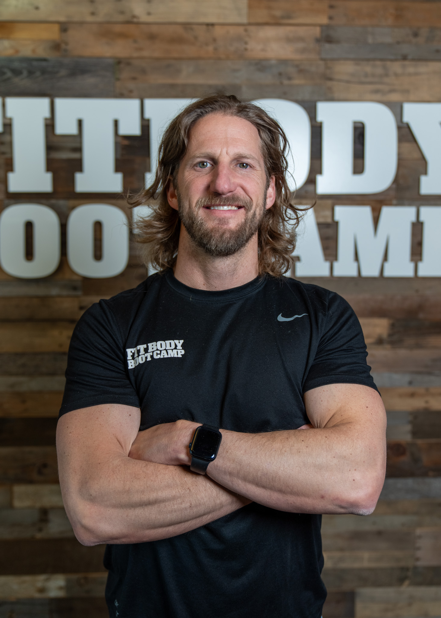 Man with long hair and beard wearing a black T-shirt and smartwatch, standing with arms crossed in front of a wooden wall with large white letters.