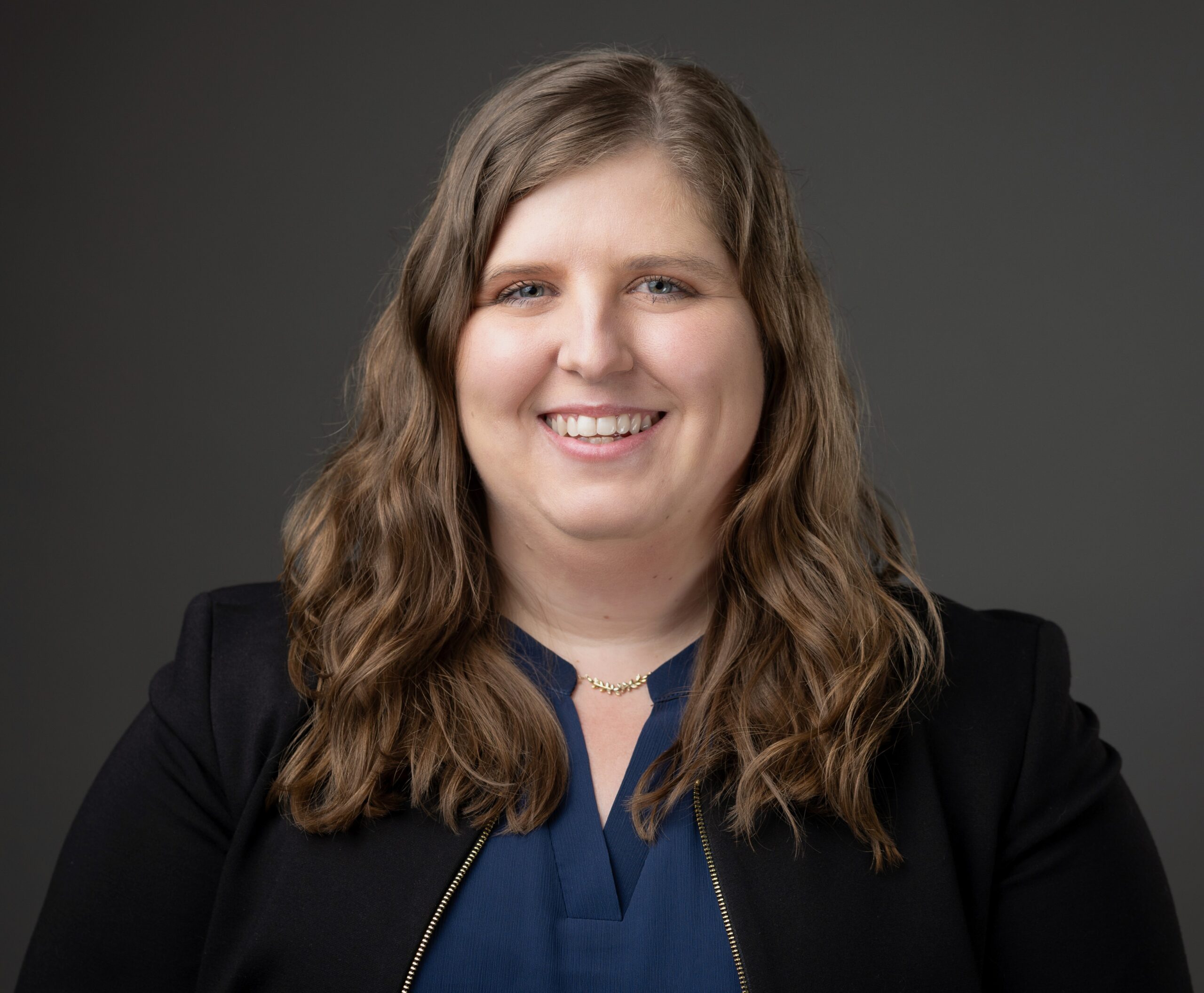 A person with long brown hair smiles, wearing a navy top and black blazer, set against a gray background.