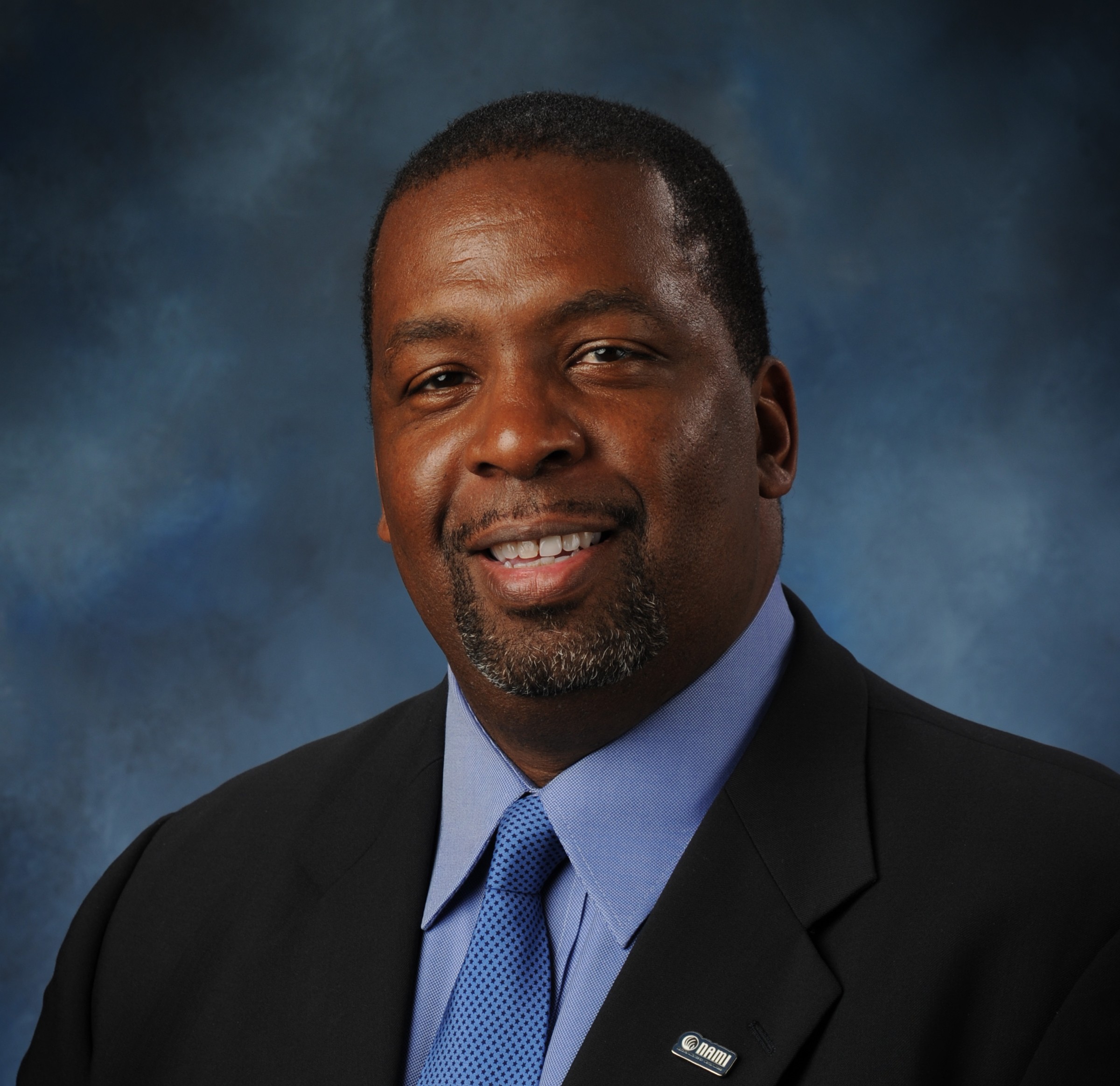 A man in a suit and tie smiles against a blue background.