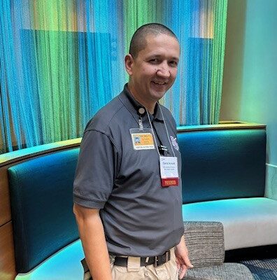 Person in a gray shirt and khaki pants stands smiling in a modern lounge with blue and green lighting in the background. They are wearing a lanyard with a badge.