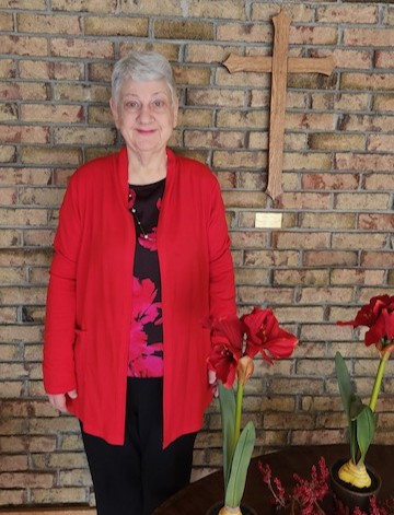 Elderly woman in a red cardigan stands next to a brick wall with a wooden cross. Red flowers in pots are on a table nearby.