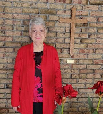 An older woman with short gray hair stands against a brick wall, wearing a red cardigan. A wooden cross hangs on the wall behind her. Two potted plants with red flowers are in the foreground.