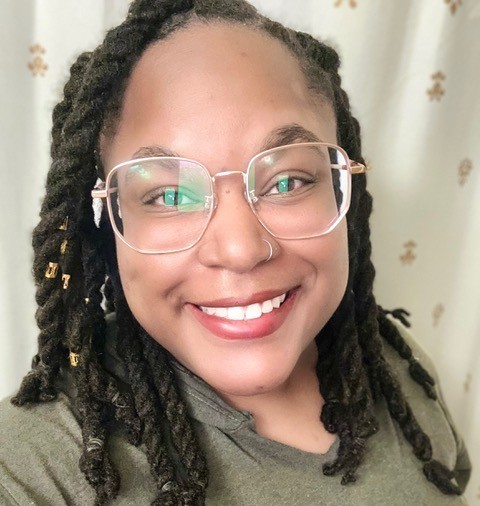 Person with glasses and braided hair smiling at the camera, standing in front of a curtain.