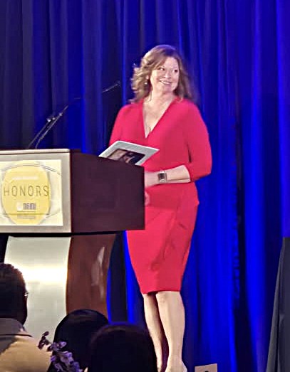 A woman in a red dress stands on stage holding papers near a podium with an "Honors" sign, with a blue curtain as background.