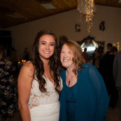 Two women smile and stand together at an indoor event with other people in the background. One woman wears a white dress, and the other wears a blue outfit.