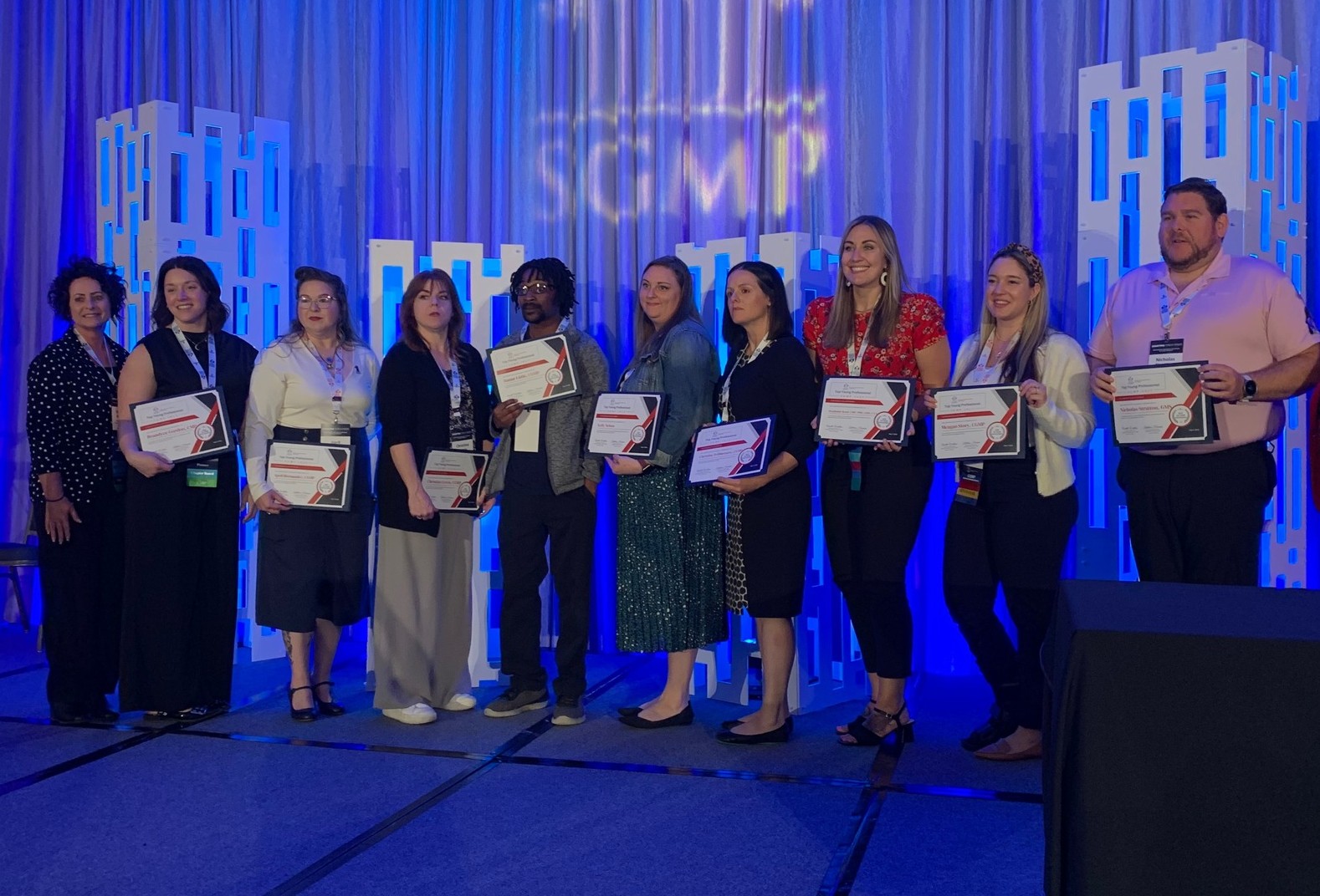 A group of ten people stand on stage holding certificates, posing for a photo at an event with a blue-lit background.