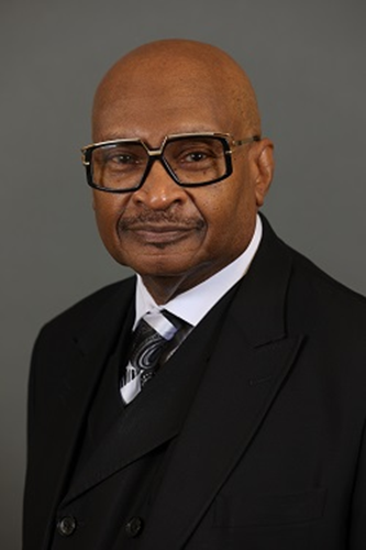 A man wearing glasses and a black suit with a patterned tie poses for a formal portrait against a plain gray background.