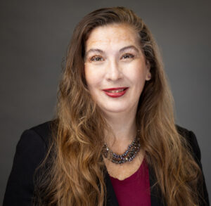 Woman with long brown hair wearing a black blazer, maroon top, and a beaded necklace, smiling at the camera against a plain dark background.
