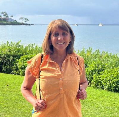 Woman in an orange shirt stands on grass near water with boats and greenery in the background under a cloudy sky.