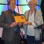 Judy Ackley presenting the Hal Madden Award to Greg Adams A man and a woman standing in front of a stage.