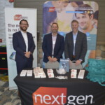 Three people stand behind a booth table with a "nextgen healthcare" banner. The table displays promotional materials.