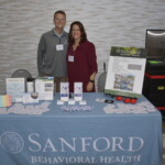 Two people stand smiling behind a Sanford Behavioral Health display table with brochures, flyers, and a poster.