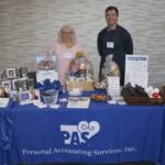 Two people standing behind a Personal Accounting Services booth with various items on display, including baskets, flyers, and promotional materials.