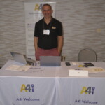 Man standing behind a display table with laptops and brochures, promoting A4i at a conference.