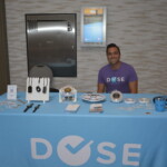 A smiling person sits at a booth with a blue tablecloth displaying Dose Health products, including wearables and promotional materials.