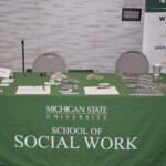 A table at a career fair features materials from Michigan State University School of Social Work, covered with a green tablecloth. Flyers, pamphlets, and small items are displayed.