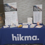 Hikma promotional booth with brochures, flyers, and two large posters about a medication. The table is covered with a blue cloth displaying the Hikma logo.