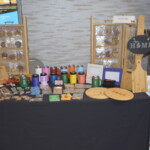 A market stall displaying various handcrafted items, including cutting boards, engraved tumblers, keychains, and wall decorations on a table with a black cloth.