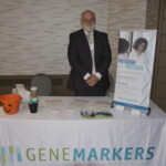 A man in a suit stands behind a table with a "Gene Markers" banner, displaying promotional materials and a Halloween-themed bucket.