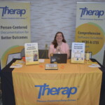 A person sits at a booth with "Therap" banners, a laptop, and informational materials displayed on the table.