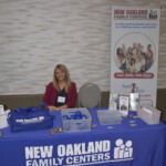 A woman sits behind a table with a New Oakland Family Centers banner, displaying informational materials and promotional items.
