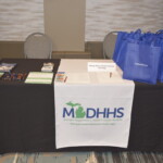 A table with a Michigan Department of Health and Human Services banner, brochures, pens, and blue tote bags.