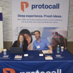 Two people seated at a table with promotional material for "protocall," featuring a backdrop with branding and text about working towards a supportive future.