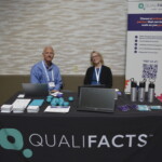 Two people seated at a Qualifacts booth with laptops, informational materials, and promotional items, including a banner displaying service details.