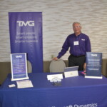 A man stands behind a table with a TMG tablecloth. There are brochures and a laptop on the table, and a TMG banner in the background.