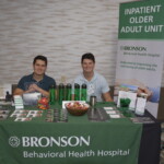 Two people sitting at a booth for Bronson Behavioral Health Hospital's Inpatient Older Adult Unit, with promotional materials and water bottles displayed on the table.