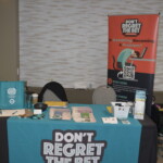 Booth with a blue table cover displaying "Don't Regret the Bet" and informational materials. A vertical banner in the background raises awareness about gambling issues.