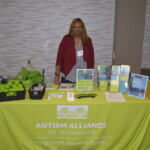 A woman stands behind an Autism Alliance of Michigan table with brochures, a basket of green apples, and various materials displayed.