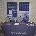 A booth for Iris Telehealth featuring promotional materials and two standing banners with illustrations and text about psychiatric care services.