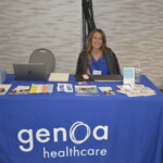 A woman sits at a table with a blue tablecloth displaying "Genoa Healthcare," surrounded by brochures, a laptop, and promotional materials.