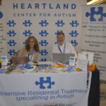 Two people sit at a table promoting the Heartland Center for Autism, surrounded by informational materials and displays about intensive residential treatment and ABA therapy.