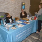 People interacting at a Behavioral Health Connections booth with informational materials displayed on a table.