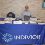 A man sits behind a table covered with a blue Indivior-branded cloth, featuring brochures and a sign.