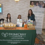 Three people smile at a table displaying brochures and snacks for StoneCrest Center, a behavioral health hospital, at an event.