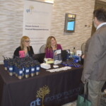 Two women sit at a booth for GT Independence, displaying promotional items. A man stands in front, engaging with them.