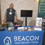 A person standing behind a table with promotional materials for Beacon Specialized Living. The table has brochures, pens, and branded items, with a backdrop featuring service highlights.
