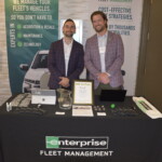 Two men standing behind a table with marketing materials and a laptop, featuring an "Enterprise Fleet Management" tablecloth. A sign promotes a chance to win AirPods.