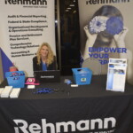 A person sits at a Rehmann booth displaying promotional materials, a laptop, and branded items. Two banners with company information are behind them.