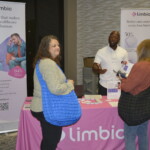 Two women talk to a man at a Limbic booth displaying banners and a tablecloth promoting clinical AI for mental healthcare.